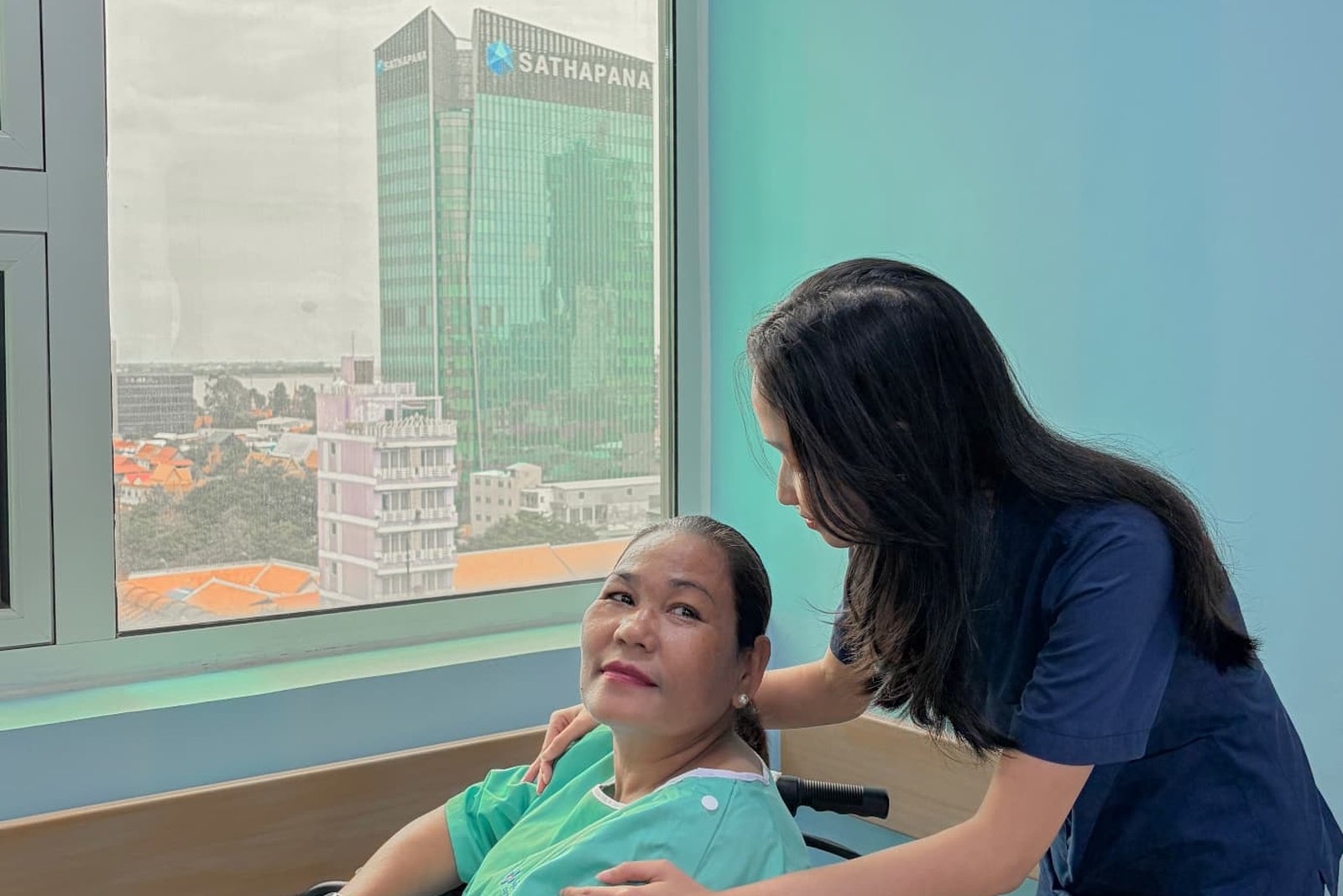 អ្នកថែទាំ Metta Lotus ស្លៀកឯកសណ្ឋានពណ៌ខៀវក្រម៉ៅ ឈរក្បែរស្ត្រីម្នាក់ដែលអង្គុយលើរទេះរុញនៅជិតបង្អួចមានពន្លឺថ្ងៃ